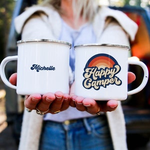 Puede incluir: Dos tazas de esmalte blanco con asas negras. Una taza dice "Michelle" y la otra taza dice "Happy Camper" con un gráfico de arcoíris.
