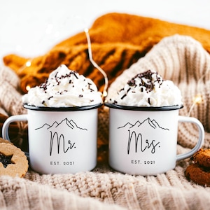 May include: Two white enamel mugs with black mountain range and text designs. The mug on the left says "Mr. EST. 2021" and the mug on the right says "Mrs. EST. 2021". The mugs are filled with whipped cream and chocolate sprinkles and are sitting on a white knitted blanket.