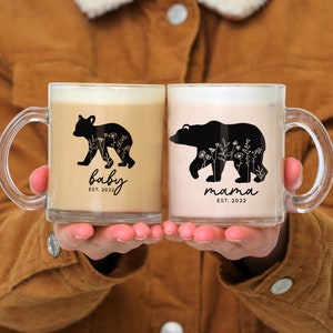 May include: Two clear glass mugs with black bear silhouettes and floral designs. The mug on the left says "baby est. 2022" and the mug on the right says "mama est. 2022".