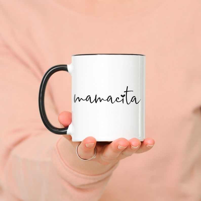 May include: White ceramic mug with a black rim and handle. The mug has the text "mamacita" written in black cursive with a small heart symbol.