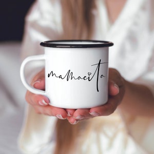 Puede incluir: Taza de esmalte blanco con borde y asa negros. La taza tiene la palabra "mamacita" escrita en cursiva negra con un peque&ntilde;o s&iacute;mbolo de coraz&oacute;n.