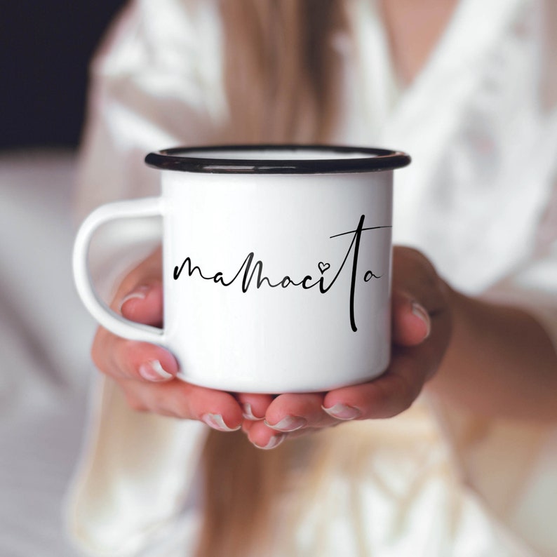 Puede incluir: Taza de esmalte blanco con borde y asa negros. La taza tiene la palabra "mamacita" escrita en cursiva negra con un peque&ntilde;o s&iacute;mbolo de coraz&oacute;n.