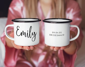 May include: Two white enamel mugs with black rims. One mug says "Emily" in black script. The other mug says "09.04.22 BRIDESMAID" in black text.
