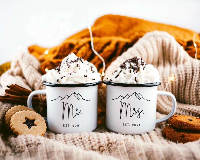 May include: Two white enamel mugs with black mountain range and text designs. The mug on the left says "Mr. EST. 2021" and the mug on the right says "Mrs. EST. 2021". The mugs are filled with whipped cream and chocolate sprinkles and are sitting on a white knitted blanket.