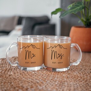 May include: Two clear glass mugs with black handles. The mugs have a mountain range design and the text "Mr. EST. 2023" and "Mrs. EST. 2023" on them. The mugs are filled with a brown liquid.