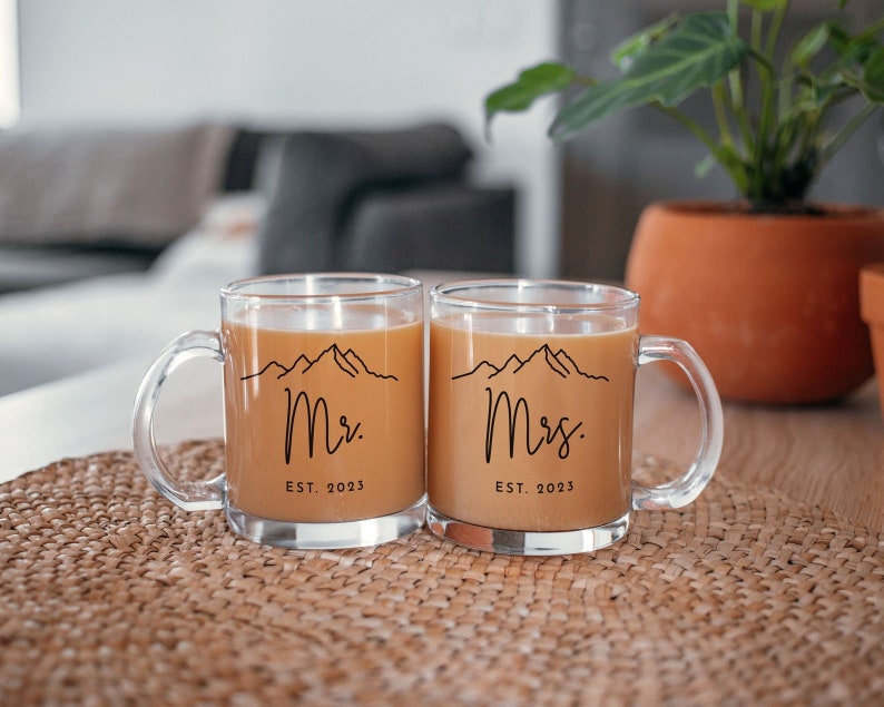 May include: Two clear glass mugs with black handles. The mugs have a mountain range design and the text "Mr. EST. 2023" and "Mrs. EST. 2023" on them. The mugs are filled with a brown liquid.