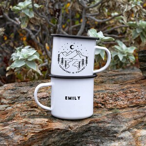 May include: Two white enamel mugs with black rims. The top mug has a black and white illustration of mountains, a river, trees, and a crescent moon with the text "Adventure is Calling". The bottom mug has the name "Emily" printed in black.