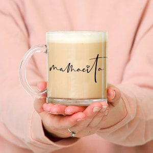 Puede incluir: Una taza de vidrio transparente con asa negra, llena de una bebida de color marr&oacute;n claro. La taza tiene la palabra "mamacita" escrita en ella en cursiva negra con un peque&ntilde;o s&iacute;mbolo de coraz&oacute;n.
