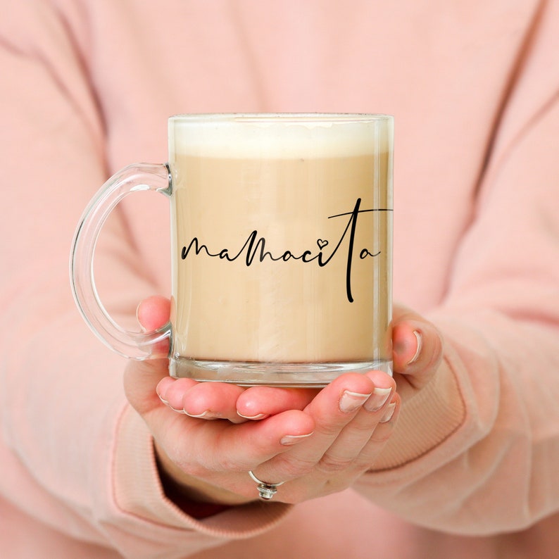 Puede incluir: Una taza de vidrio transparente con asa negra, llena de una bebida de color marr&oacute;n claro. La taza tiene la palabra "mamacita" escrita en ella en cursiva negra con un peque&ntilde;o s&iacute;mbolo de coraz&oacute;n.