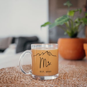 May include: A clear glass mug with a black outline of mountains and the text "Mr. EST. 2023" filled with a light brown beverage.