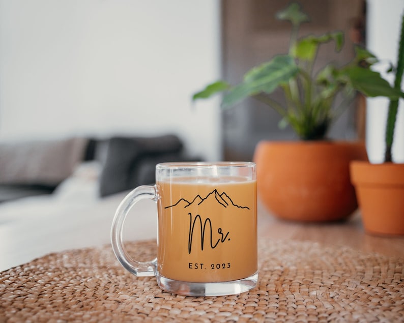May include: A clear glass mug with a black outline of mountains and the text "Mr. EST. 2023" filled with a light brown beverage.