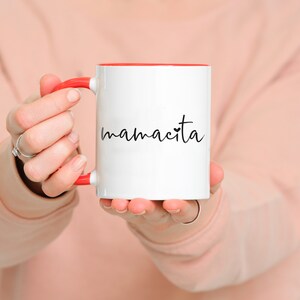 May include: White ceramic mug with a red rim and handle. The mug has the word "mamacita" written on it in black cursive with a small heart.