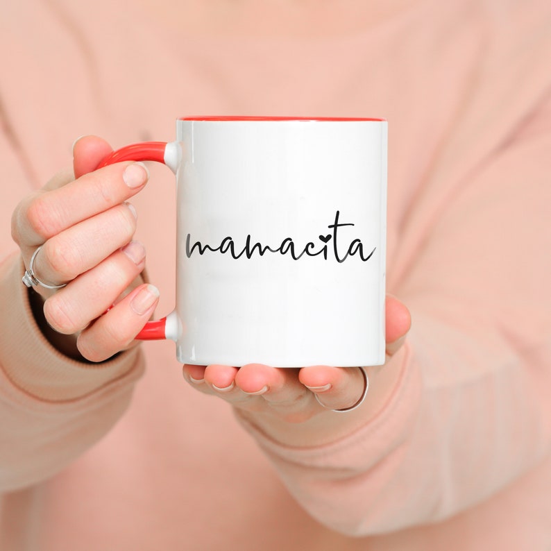 May include: White ceramic mug with a red rim and handle. The mug has the word "mamacita" written on it in black cursive with a small heart.
