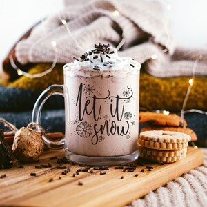 May include: A glass mug with a white and brown beverage inside. The mug has a black design that says "Let it snow" with snowflakes. The mug is sitting on a wooden surface with chocolate sprinkles scattered around it.