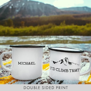 Puede incluir: Dos tazas de esmalte blanco con bordes negros. Una taza dice "MICHAEL" y la otra taza dice "I'D CLIMB THAT" con una silueta de montaña. Las tazas están sentadas en una manta a rayas amarillas y blancas frente a un fondo borroso de un río y montañas.