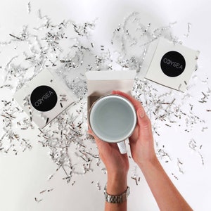 May include: White ceramic mug with a white logo that says "ODYSSEA" on it. The mug is sitting on a white surface with silver confetti.