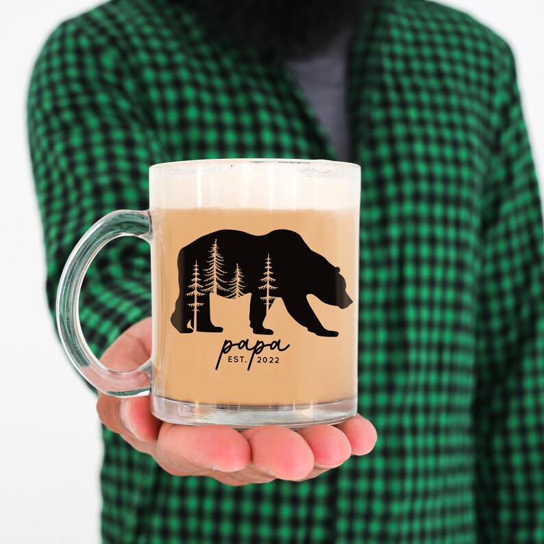 May include: A clear glass mug with a black bear silhouette and trees inside the bear. The mug has the text "papa est. 2022" printed on it.