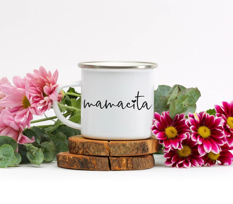 May include: White enamel mug with a black inscription that reads "mamacita" with a small heart symbol. The mug is sitting on a wooden coaster.