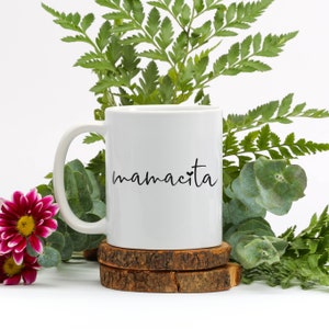 May include: White ceramic mug with a black inscription that reads "mamacita" with a small heart symbol. The mug is sitting on a wooden coaster.