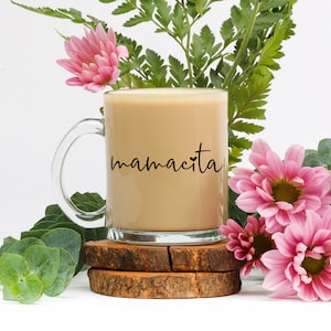 May include: A glass coffee mug with a black handle and the text "mamacita" printed on the side. The mug is filled with coffee and is sitting on a wooden coaster.