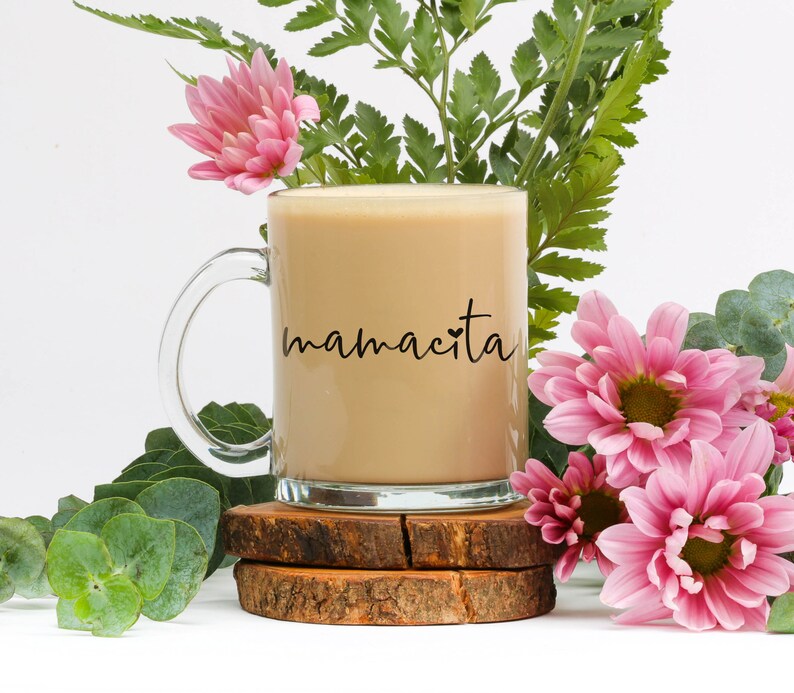 May include: A glass coffee mug with a black handle and the text "mamacita" printed on the side. The mug is filled with coffee and is sitting on a wooden coaster.