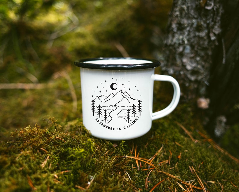 May include: White enamel camping mug with a black rim and handle. The mug has a black and white illustration of mountains, a crescent moon, and trees with the text "Adventure is Calling".