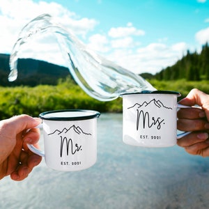 May include: Two white enamel mugs with black rims and handles. The mugs have a black mountain range graphic and the text "Mr. EST. 2021" and "Mrs. EST. 2021". The mugs are being clinked together with water splashing in the air.