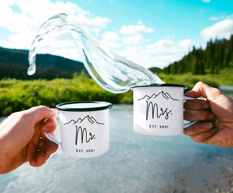 May include: Two white enamel mugs with black rims and handles. The mugs have a black mountain range graphic and the text "Mr. EST. 2021" and "Mrs. EST. 2021". The mugs are being clinked together with water splashing in the air.
