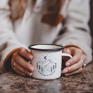 May include: White enamel mug with a black rim and handle. The mug has a black and white illustration of mountains, a crescent moon, and stars with the text "Adventure is Calling".