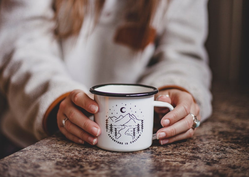 May include: White enamel mug with a black rim and handle. The mug has a black and white illustration of mountains, a crescent moon, and stars with the text "Adventure is Calling".