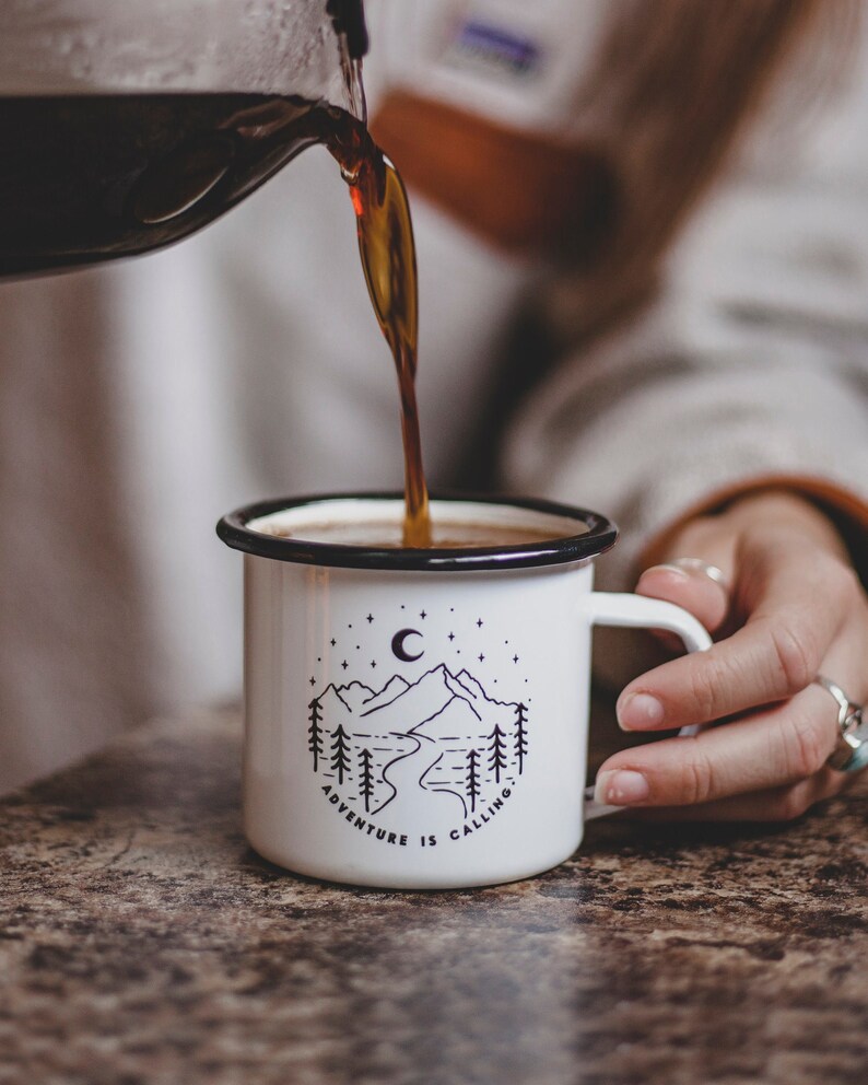 May include: White enamel mug with a black rim and handle. The mug has a black and white illustration of a mountain range, a crescent moon, and stars. The text "Adventure is Calling" is printed on the mug.