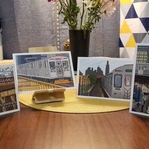 May include: Four art prints of Chicago train scenes. Each print features a different perspective of the city's elevated train system, with detailed illustrations of trains, tracks, and urban landscapes. The prints are displayed on a wooden table.
