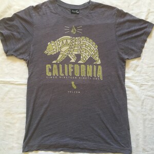 May include: Gray t-shirt with a yellow graphic of a bear with a geometric pattern and the text "CALIFORNIA SINCE NINETEEN NINETY ONE" and a small graphic of the state of California with the text "VOLCOM" underneath.