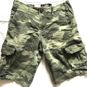 Puede incluir: Shorts cargo de camuflaje verde y marrón con un logotipo de Hollister en la parte delantera. Los pantalones cortos tienen varios bolsillos y un cierre de botón.