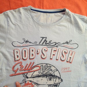 以下が含まれることがあります： 薄い青色のTシャツに、魚、フライドポテト、そして「The Cool Guaranteed Bob's Fish Grill 1947」というテキストが書かれたヴィンテージスタイルのグラフィックデザインが施されています。