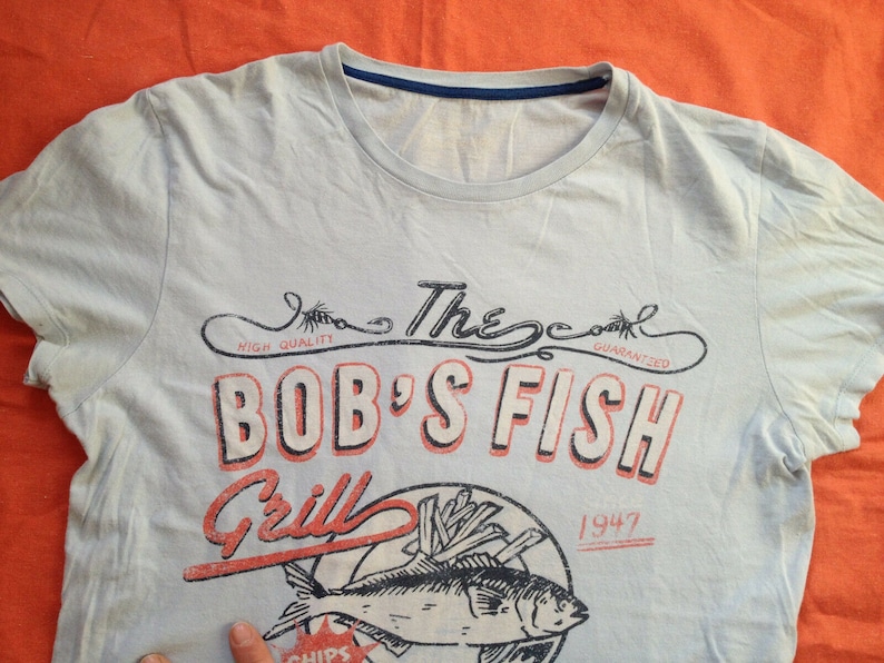 以下が含まれることがあります： 薄い青色のTシャツに、魚、フライドポテト、そして「The Cool Guaranteed Bob's Fish Grill 1947」というテキストが書かれたヴィンテージスタイルのグラフィックデザインが施されています。