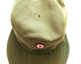 May include: Olive green military-style cap with a rounded crown and a short, curved brim. The cap features a circular emblem with red, white, and black details. The fabric appears to be a durable, woven material. There is a small hole on the top of the cap.