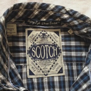 May include: Close-up of a blue, white, and black plaid shirt. The shirt has a collar and a label with the word "SCOTCH" in a diamond shape. The text "Enjoy Life Wear Scotch" is above the label.