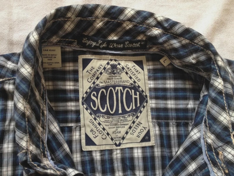 May include: Close-up of a blue, white, and black plaid shirt. The shirt has a collar and a label with the word "SCOTCH" in a diamond shape. The text "Enjoy Life Wear Scotch" is above the label.