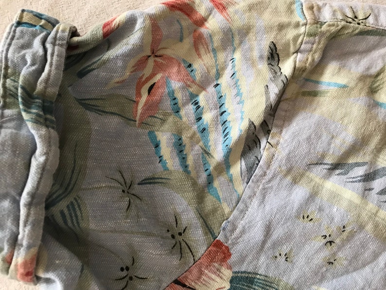 May include: A light blue shirt with a floral pattern. The pattern features pink, blue, and green flowers and leaves. The shirt has a button-up collar and a long sleeve.