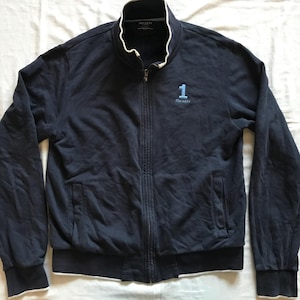Puede incluir: Una chaqueta azul marino con cremallera, cuello blanco y ribetes blancos alrededor de los puños. La chaqueta tiene un solo bolsillo en el lado izquierdo y un logotipo en el pecho que dice "1 Hackett".