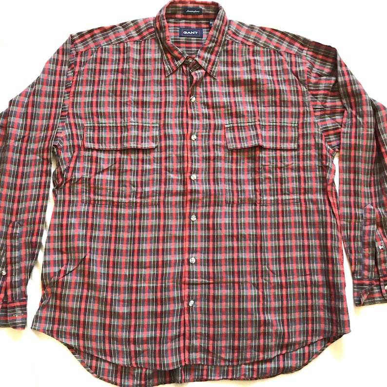 Puede incluir: Una camisa de manga larga con un estampado de cuadros rojos, negros y verdes. Tiene dos bolsillos en el pecho y un cuello abotonado. La marca "GANT" es visible en el cuello.