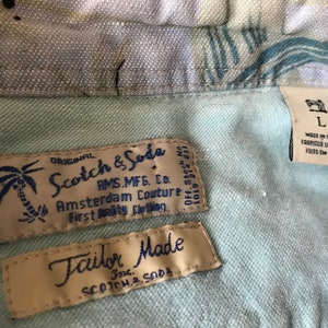 May include: A close-up of two labels on a light blue shirt. The top label is white with black text that reads "ORIGINAL Scotch & Soda AMS.MFG. Co. Amsterdam Couture First Quality Clothing Off. Stock No. 1015 851211". The bottom label is beige with blue text that reads "Tailor Made Inc. SCOTCH & SODA".
