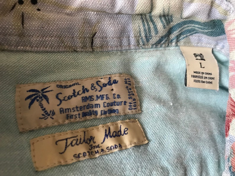 May include: A close-up of two labels on a light blue shirt. The top label is white with black text that reads "ORIGINAL Scotch & Soda AMS.MFG. Co. Amsterdam Couture First Quality Clothing Off. Stock No. 1015 851211". The bottom label is beige with blue text that reads "Tailor Made Inc. SCOTCH & SODA".