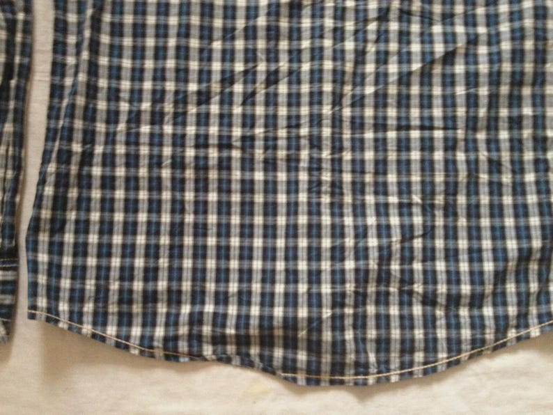 May include: Close-up of a blue, white, and black plaid shirt. The fabric is a woven pattern of small squares. The shirt has long sleeves and a curved hemline. The shirt is made of a soft, comfortable material.