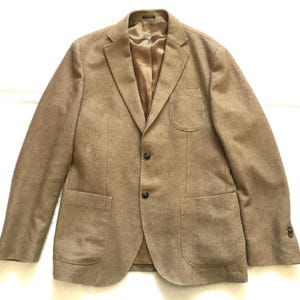 May include: A tan blazer with a two-button closure, two front pockets, and a chest pocket. The blazer has a notched lapel and a brown lining. The sleeves have three buttons at the cuff.