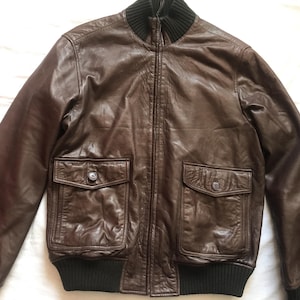 May include: A brown leather bomber jacket with a black ribbed collar, cuffs, and hem. The jacket features two front pockets with button closures. The leather has a slightly distressed appearance.