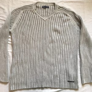 Puede incluir: Un suéter de punto acanalado gris claro con cuello en V. El suéter de manga larga tiene canalé vertical y una pequeña etiqueta cerca del dobladillo inferior. El suéter está hecho de un material suave y cómodo.