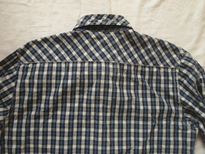 May include: Back view of a blue, black, and white plaid button-down shirt. The shirt has a collar and a yoke detail. The fabric is a woven cotton or similar material. The shirt is likely a casual wear item.