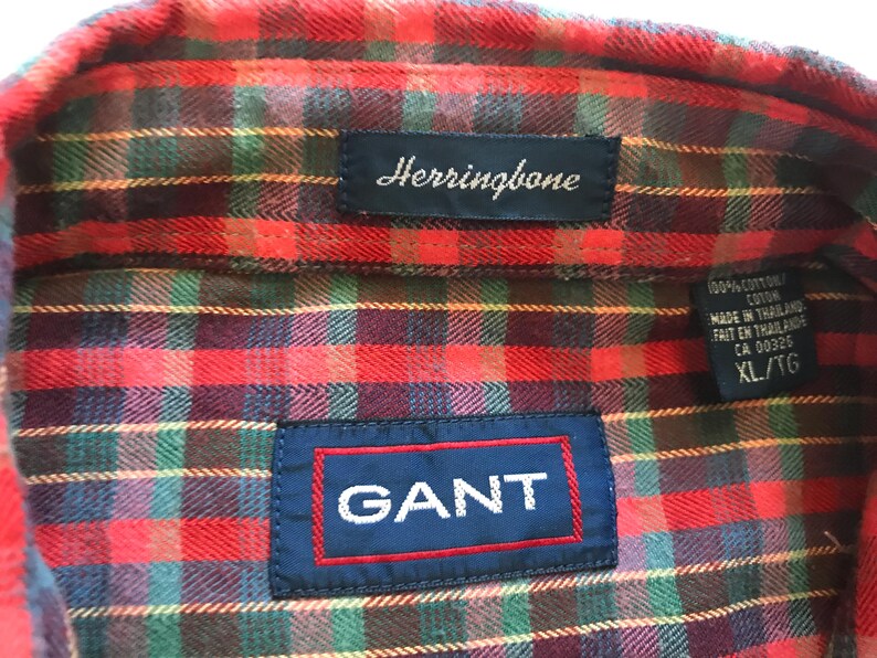 Puede incluir: Primer plano de una camisa a cuadros roja, verde y azul con la marca "GANT" en un parche azul. La camisa tambi&eacute;n tiene una etiqueta "Herringbone" y una etiqueta que indica XL/TG.